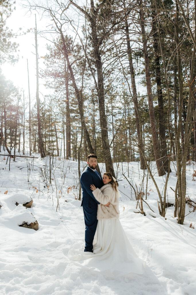 Bride and groom standing in the snow in tall trees with the sun shining through the trees