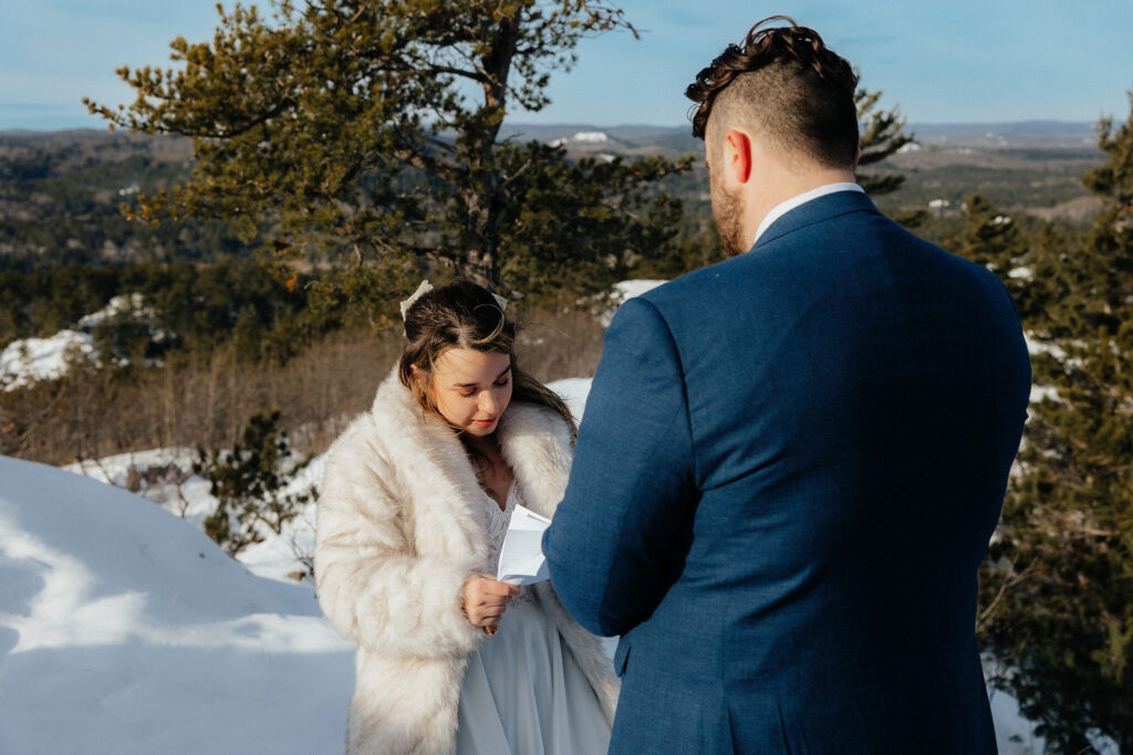 bride and groom reading their vows to each other. blue sky, trees and snow in the background